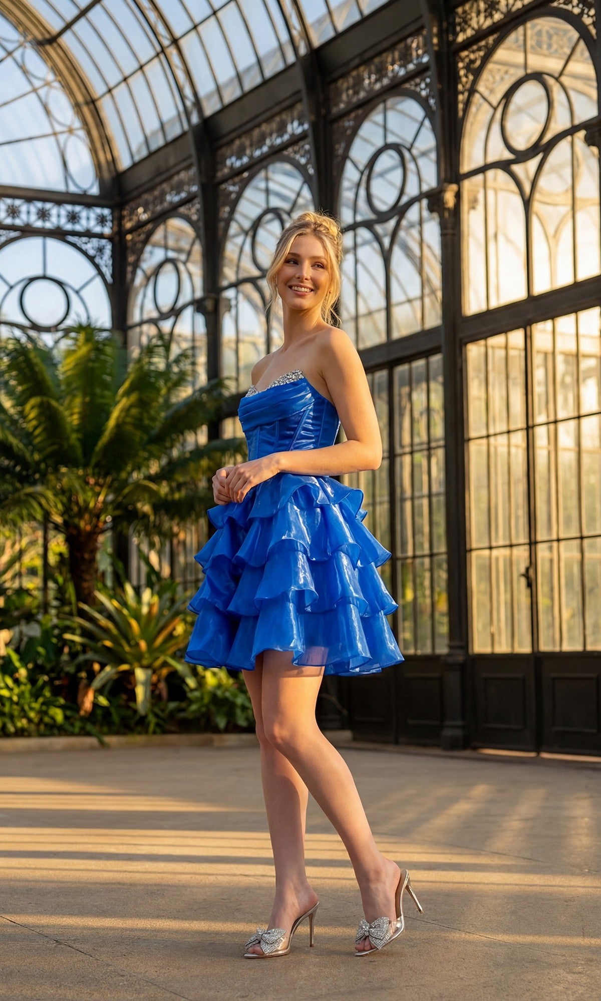 Wearing the Juliet JT7134S short homecoming dress—a vibrant blue, strapless organza design with a ruffle a-line mini skirt—a woman stands smiling in a glass conservatory, surrounded by plants and sunlight streaming through the large windows, her look completed with silver heels.